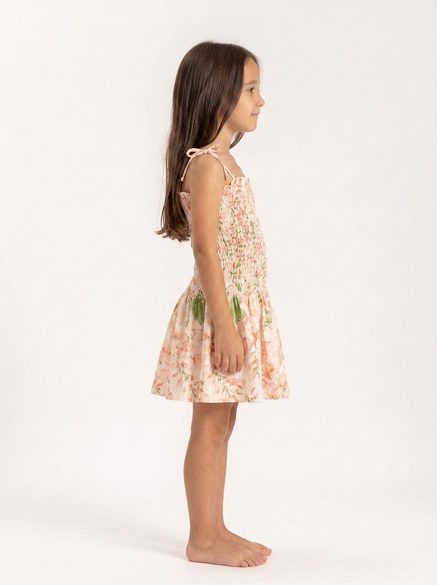 A young girl with long brown hair is shown in profile wearing a sleeveless, smocked floral dress with thin shoulder ties. She is standing barefoot against a clean, off-white studio background.