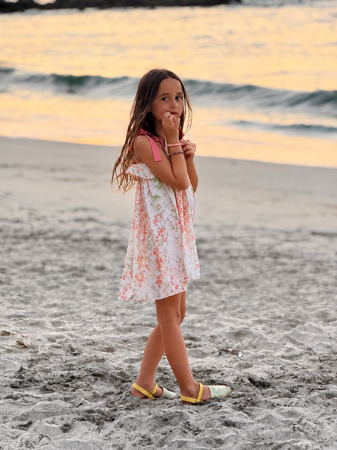 A young girl with long brown hair stands on a sandy beach at sunset, wearing a coral floral dress with pink and green accents and yellow sandals.