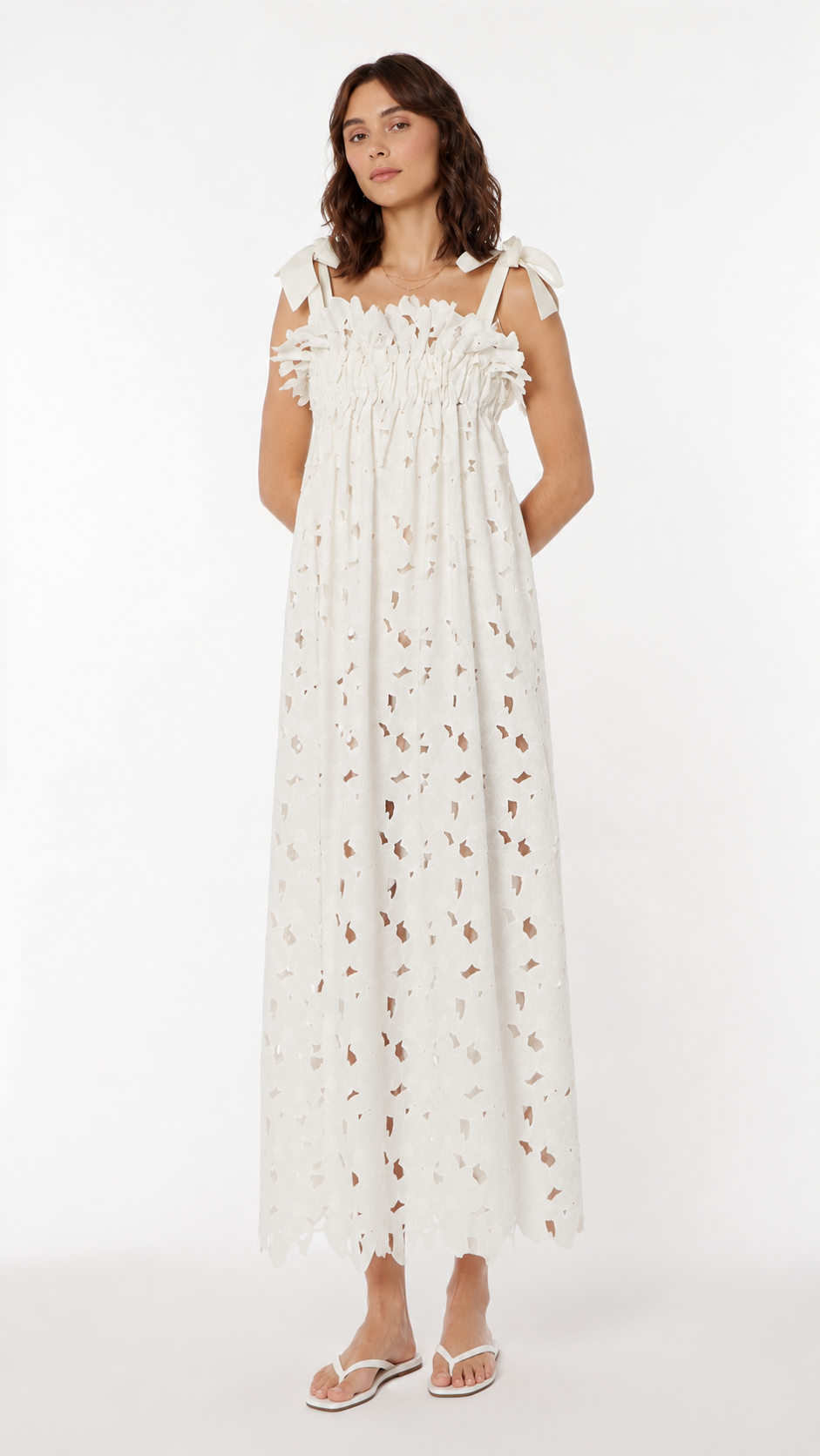 A woman modeling a loose-fitting white cotton lace maxi dress with tie-shoulder, paired with white sandals, against a plain white studio background.