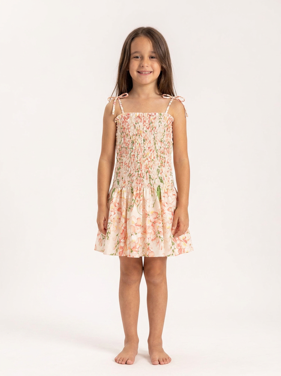 A young girl with long brown hair stands barefoot against a plain white studio background, wearing a sleeveless smocked dress with thin shoulder ties and a pink floral print.